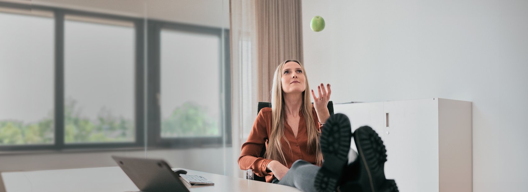 Frau entspannt mit hochgelegten Füßen auf einem Tisch, während sie einen Apfel in die Luft wirft, symbolisiert den Genuss von Obst in Tüten.
